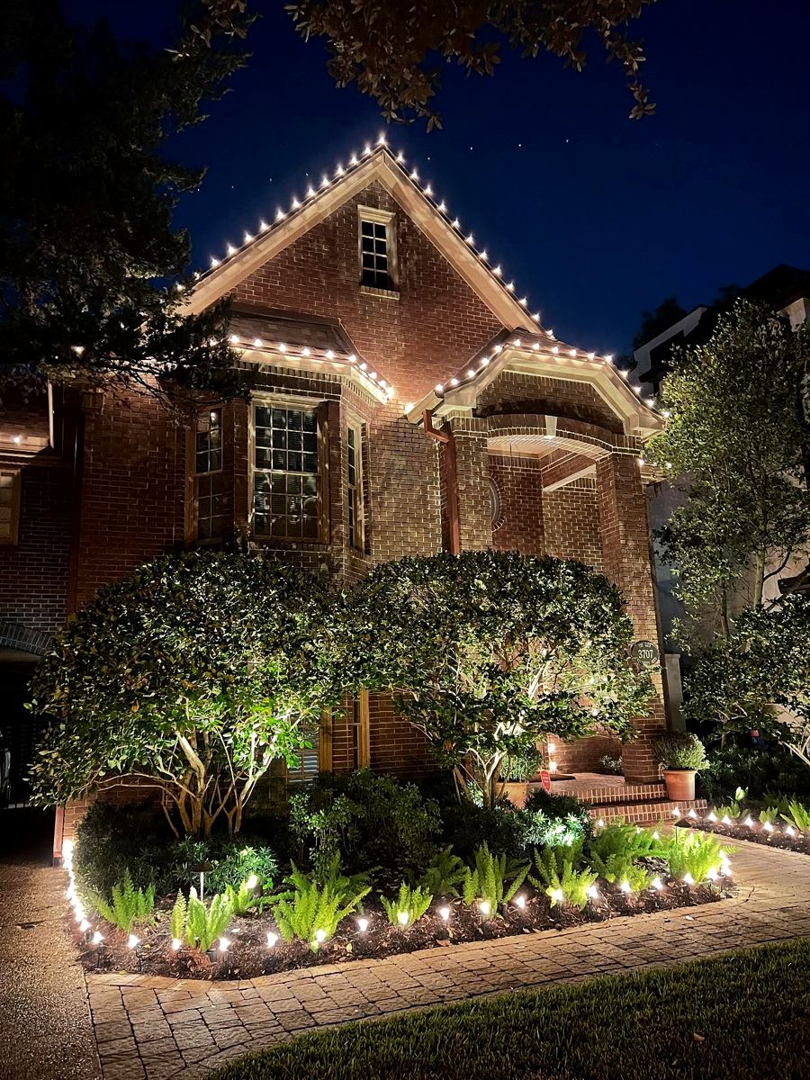 Brick home with warm white bulbs outlining steep gables above a lit garden bed