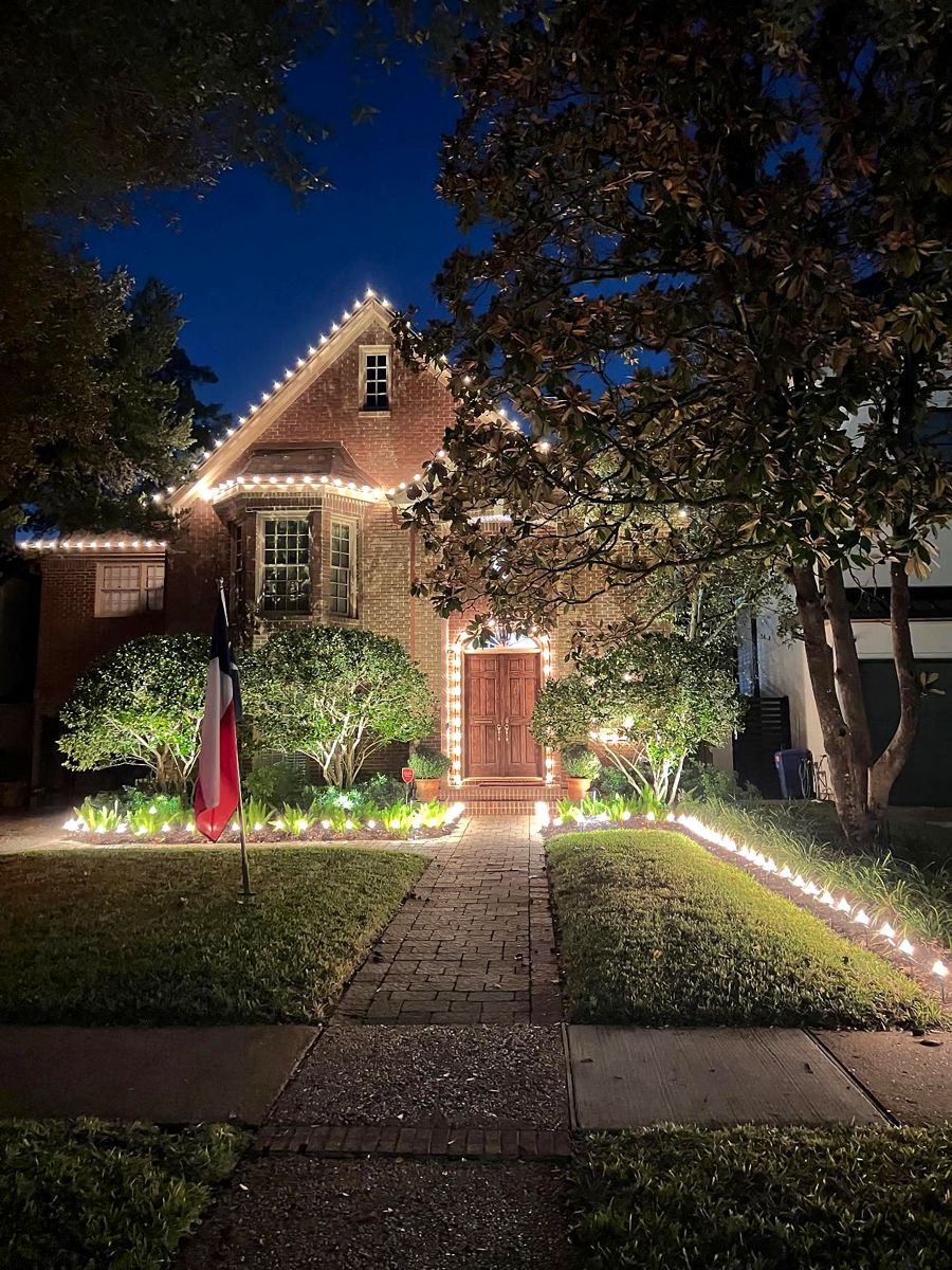 Brick cottage with warm white roofline bulbs, a lit front walkway and a Texas flag