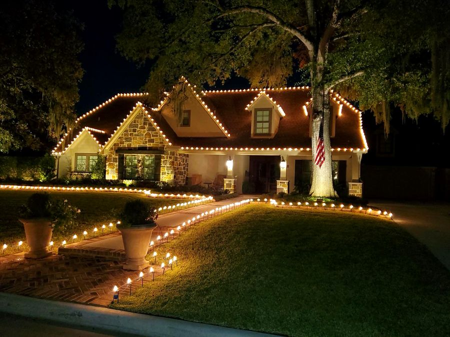 Stone-front home with warm white bulbs on every gable, a lit path and a flag on the oak tree