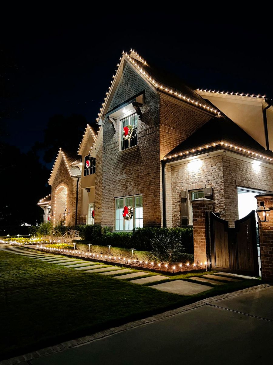 Brick two-story with warm white bulbs along the gables, wreaths in the windows and a lit walkway